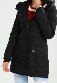 Svart parkajacka med en huva fodrad med päls, dragkedja och knäppningar, dragsko i midjan och två framfickor. Strukturerad tyg.