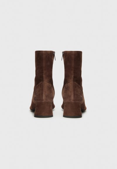 Botas de ante marrón hasta el tobillo con un tacón cuadrado, que presentan una textura suave y un talón cerrado, con un detalle de cremallera en la parte superior.