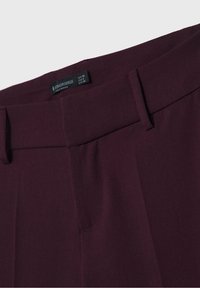 Burgundowe spodnie o gładkiej tkaninie, prostym kroju i bocznych kieszeniach. Widoczna metka z informacjami o rozmiarze.