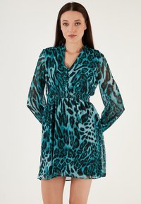 Haljina s leopard printom u tirkiznoj i crnoj boji, izrađena od prozirnog materijala. Ima v-izrez, duge rukave i elastičnu strukicu za prilagođen siluetu.