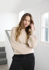 Beige sweater with ribbed balloon sleeves and round neckline, paired with black pants featuring a drawstring. Minimalist backdrop.