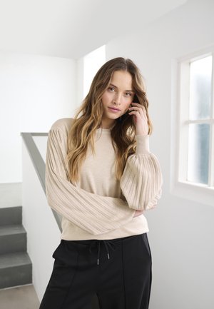 Beige sweater with ribbed balloon sleeves and round neckline, paired with black pants featuring a drawstring. Minimalist backdrop.