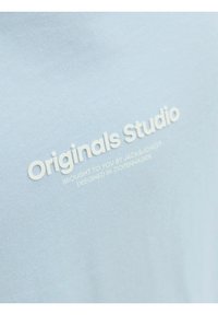 Světle modré bavlněné tričko s bílým embošovaným logem "Originals Studio" s dalším textem níže. Hladký povrch.