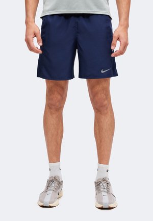 Homme portant un short de sport bleu marine, des chaussettes blanches et des chaussures de course Nike beiges, debout devant un fond uni.