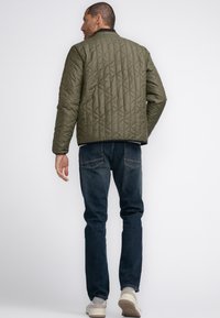 Olivgrüne gesteppte Jacke mit schwarzem Kragen, die ein Rautenmuster aufweist, kombiniert mit dunkelblauen Jeans und hellen Sneakers.