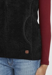 Svart fleeceväst med dragkedja, två sidfickor och en brun logotypbadge. Under västen syns en ribbad maroon långärmad tröja.