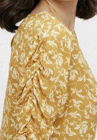Blouse jaune moutarde avec un motif floral, tissu texturé et détails plissés sur les manches. Encolure ronde et coupe décontractée.