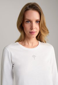 White long-sleeve t-shirt with a round neck and a subtle rhinestone floral design at the center front. Soft cotton material.