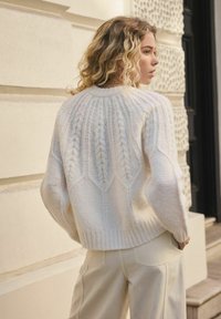 Maglione bianco lavorato a maglia con design a trecce testurizzate, maniche ampie e orlo a coste. Abbinato a pantaloni chiari a vita alta.