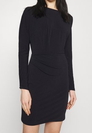 Femme portant une robe noire cintrée à manches longues avec un détail drapé à la taille sur un fond blanc uni.