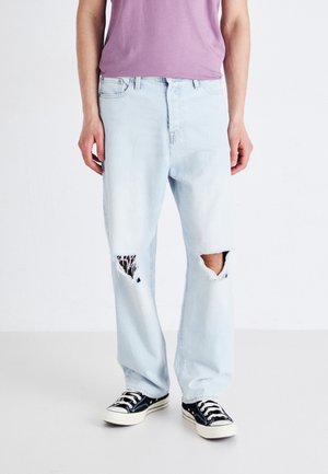 Person trägt hellblaue zerrissene Jeans mit Löchern an den Knien, schwarz-weiße Sneakers und ein lila Hemd, steht vor weißem Hintergrund.