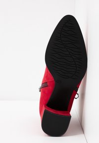 Bottine en daim rouge avec semelle en caoutchouc noire, talon bloc et fermeture éclair latérale, vue par le dessous sur fond blanc.