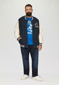 Navy and cream varsity jacket with white stripes, over a blue graphic tee, paired with dark jeans and white sneakers.