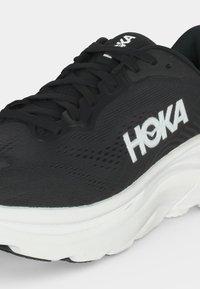 Μαύρο παπούτσι για τρέξιμο HOKA με λευκό λογότυπο, μαύρα κορδόνια και χοντρή λευκή σόλα, εμφανιζόμενο από εμπρόσθια πλευρική γωνία.