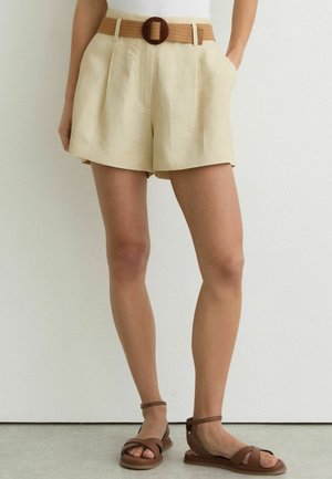 Femeie purtând pantaloni scurți plisati bej cu talie înaltă, cu centură împletită și sandale maro cu barete la gleznă, stând în fața unui perete alb.