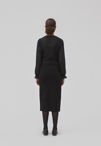 Haut à manches longues noir avec un design texturé, associé à une jupe midi noire ajustée et des chaussures noires. Fini mat, détails minimaux.