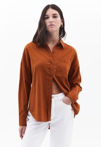 Rost-orange Hemd mit Knöpfen, einer Brusttasche, langen Ärmeln und einem abgerundeten Saum, kombiniert mit weißen Hosen. Glatte Textur, lässige Passform.