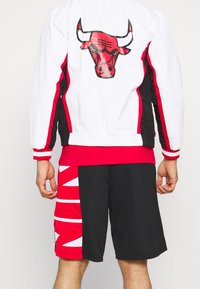 Hvid og sort jakke med røde detaljer, der har et tyre-motiv på ryggen. Lagdelt rød skjorte under sorte shorts med vinkelret rødt og hvidt design.