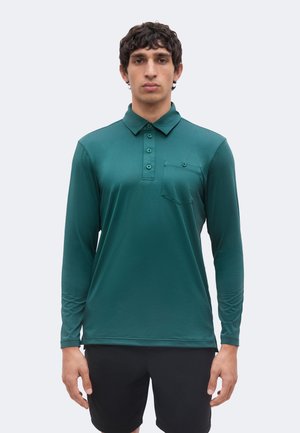 Homme portant un polo à manches longues bleu sarcelle avec boutons et poche poitrine, assorti à un short noir, debout devant un fond uni.