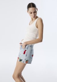 Shorts in denim con lavaggio blu, caratterizzati da ricami colorati di frutta e bevande sulle tasche. Abbinati a un top a canotta bianco a coste.