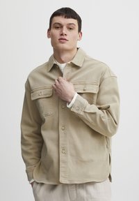 Light beige denim shirt with two chest pockets, button front, and rounded hem. Loose fit with long sleeves and subtle texture.