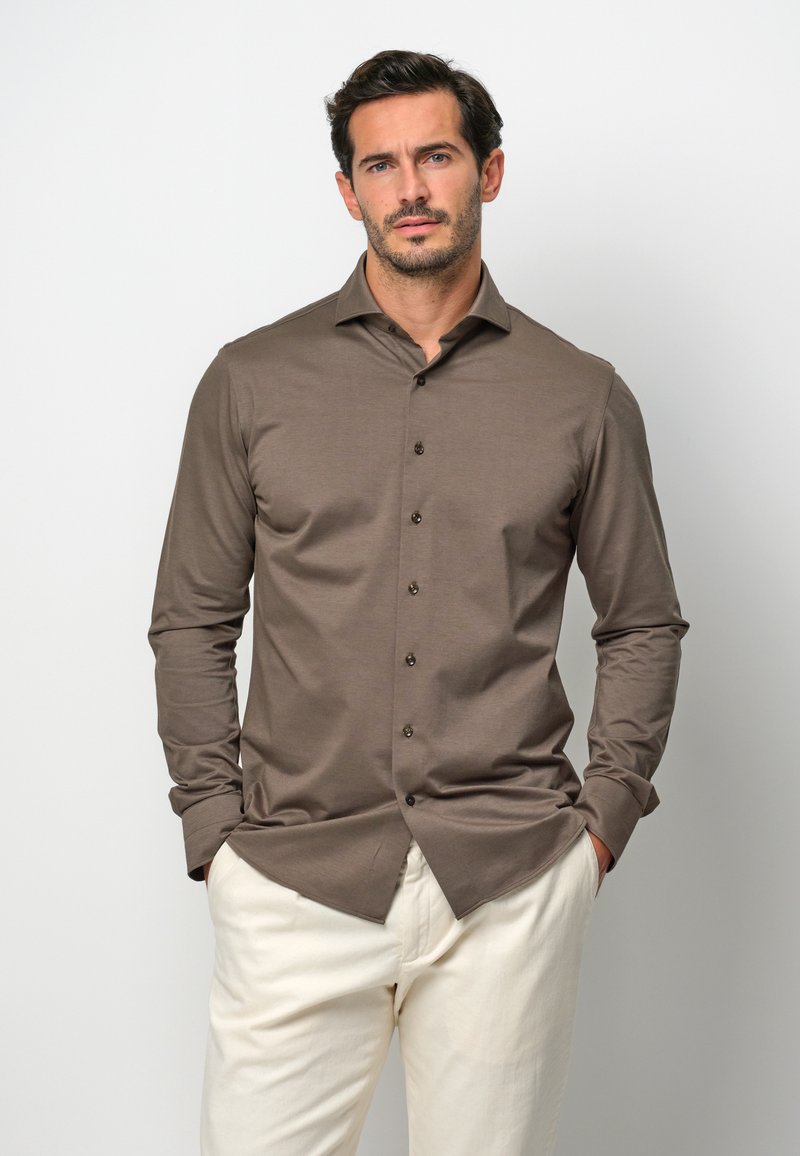 Camicia marrone a maniche lunghe con colletto classico e chiusura con bottoni. Il tessuto appare liscio con una leggera lucentezza. Indossata con pantaloni di colore chiaro.