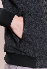 Giacca nera con zip e un motivo maculato. Presenta polsini e orlo neri solidi, con una tasca frontale e dettagli della zip lisci.