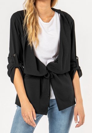 Giacca nera leggera con design a nodo davanti, maniche arrotolate e vestibilità morbida. Indossata su una maglietta bianca e jeans blu.