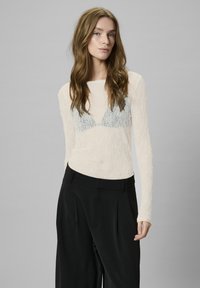 Light, textured long-sleeve top in cream with a sheer lace pattern and a blue lace triangle design beneath. Paired with black high-waisted trousers.