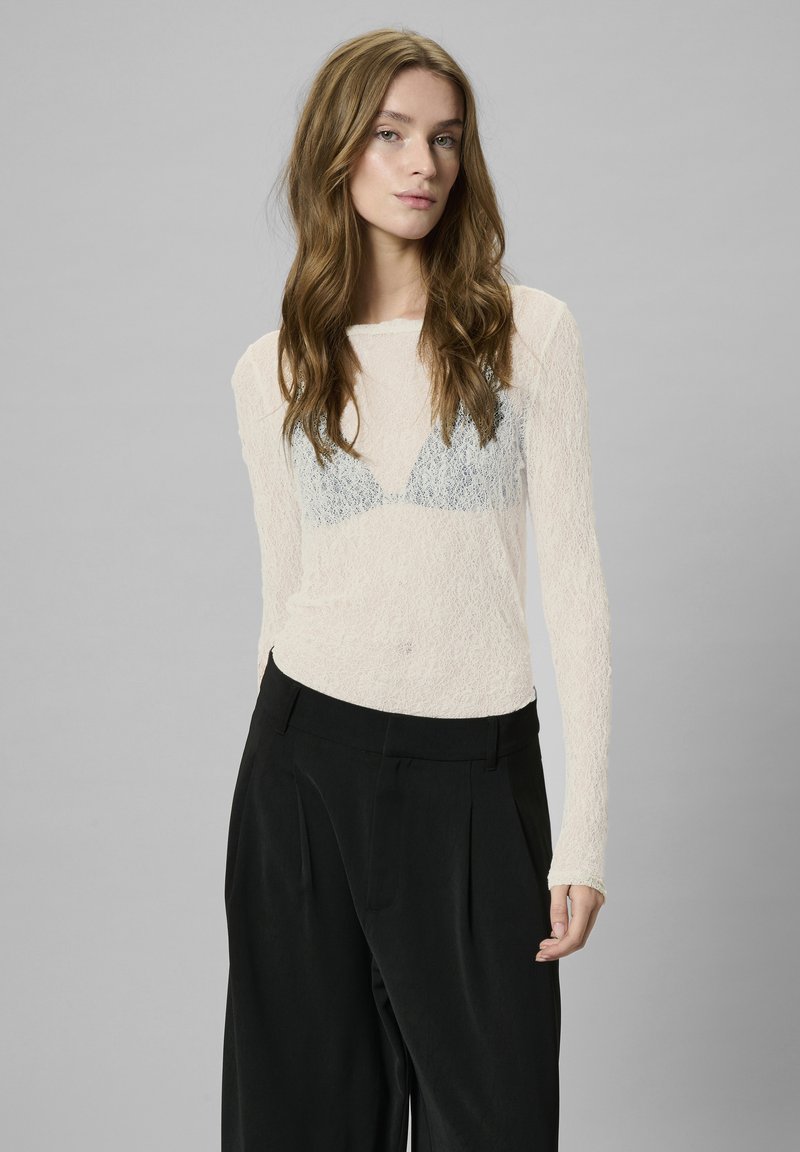 Light, textured long-sleeve top in cream with a sheer lace pattern and a blue lace triangle design beneath. Paired with black high-waisted trousers.