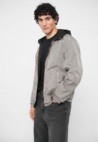 Jaunas vyras su garbanotais plaukais, vilkintis raštuotą užtrauktuku užsegamą striukę su gobtuvu, po kuria dėvi juodą marškinį ir tamsiai pilkas džinsines kelnes, stovi su viena ranka kišenėje.