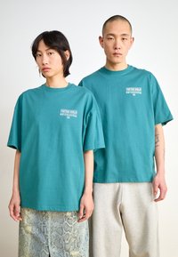 Teal, oversized cotton t-shirts with short sleeves, featuring a white logo on the left chest, worn by two models with casual bottoms.