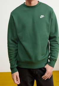 Grön sweatshirt med rundad halsringning, ribbade ärmslut och nederkant. Har en vit Nike-logotyp på bröstet. Tyget verkar mjukt och strukturerat.