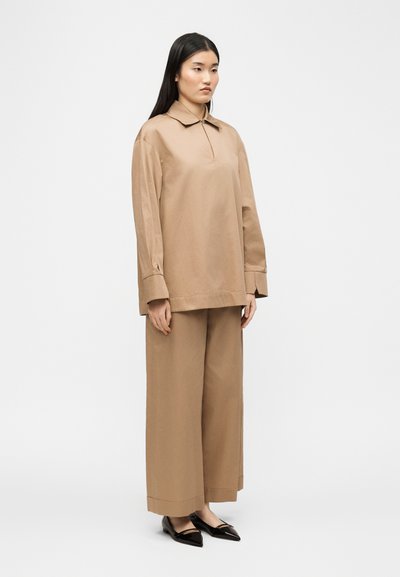 Femme debout portant un haut beige à manches longues avec col et un pantalon beige à jambes larges, chaussée de ballerines noires à bout pointu, sur un fond blanc uni.