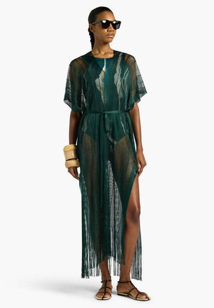 Donna che indossa un copricostume trasparente verde scuro con frange sopra il costume da bagno, occhiali da sole neri, braccialetti color bronzo e sandali neri, in piedi davanti a uno sfondo bianco.