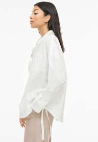 White, oversized shirt made of lightweight fabric. Features long sleeves, a chest pocket, and ties at the side. Worn with light-colored pants.