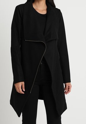 Mujer con un abrigo largo negro asimétrico con cremallera, pantalones negros y una camisa negra, de pie frente a un fondo claro y liso.