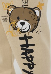 Beżowe spodnie dresowe mają drukowany motyw misia w kolorze brązowym, z czarnymi i złotymi akcentami oraz wyraźnym czarnym napisem "Szczęśliwy".