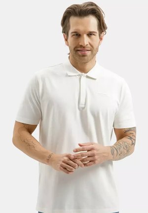 Mężczyzna o jasnobrązowych włosach, ubrany w białą koszulkę polo z krótkim rękawem, pokazujący tatuaże na obu przedramionach, na jasnym, jednolitym tle.