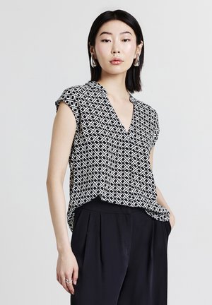 Donna con capelli neri lisci che indossa una blusa a fantasia bianca e nera e pantaloni neri, in posa con una mano in tasca.