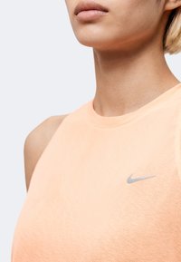 Κοντινό πλάνο ενός ατόμου που φοράει αμάνικο αθλητικό τοπ ροδακινί με μικρό γκρι λογότυπο Nike στο στήθος, με φόντο απλό και φωτεινό.