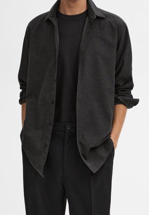 Homme portant une chemise noire côtelée boutonnée par-dessus un t-shirt noir uni avec les manches retroussées et un pantalon noir, les mains dans les poches.
