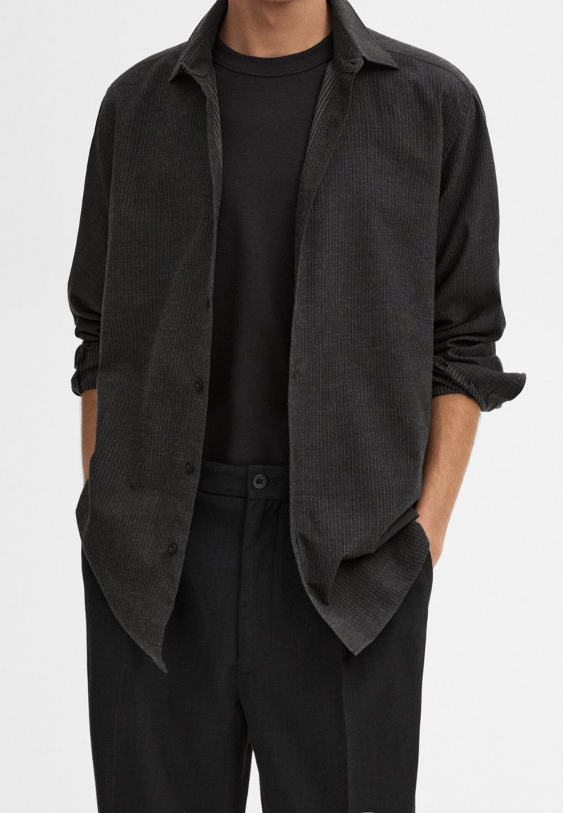 Homme portant une chemise noire côtelée boutonnée par-dessus un t-shirt noir uni avec les manches retroussées et un pantalon noir, les mains dans les poches.
