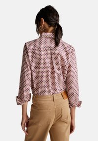 Langärmlige Bluse mit lila und cremefarbenem geometrischem Muster, weichem Stoff, Kragen und aufgerollten Ärmeln, kombiniert mit beigen, eng anliegenden Hosen.