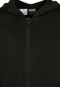 Juodas užtrauktuku užsegamas džemperis, pagamintas iš minkštos medžiagos, su priekiniu užtrauktuku ir prisegamu gobtuvu. Matoma etiketė ant vidinio kaklo.