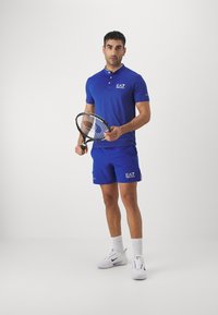 Mėlynas polo ir šortų komplektas, pagamintas iš kvėpuojančios medžiagos. Yra mygtukai, siuvinėtas logotipas, dėvimas su baltais sportiniais bateliais ir šortais.