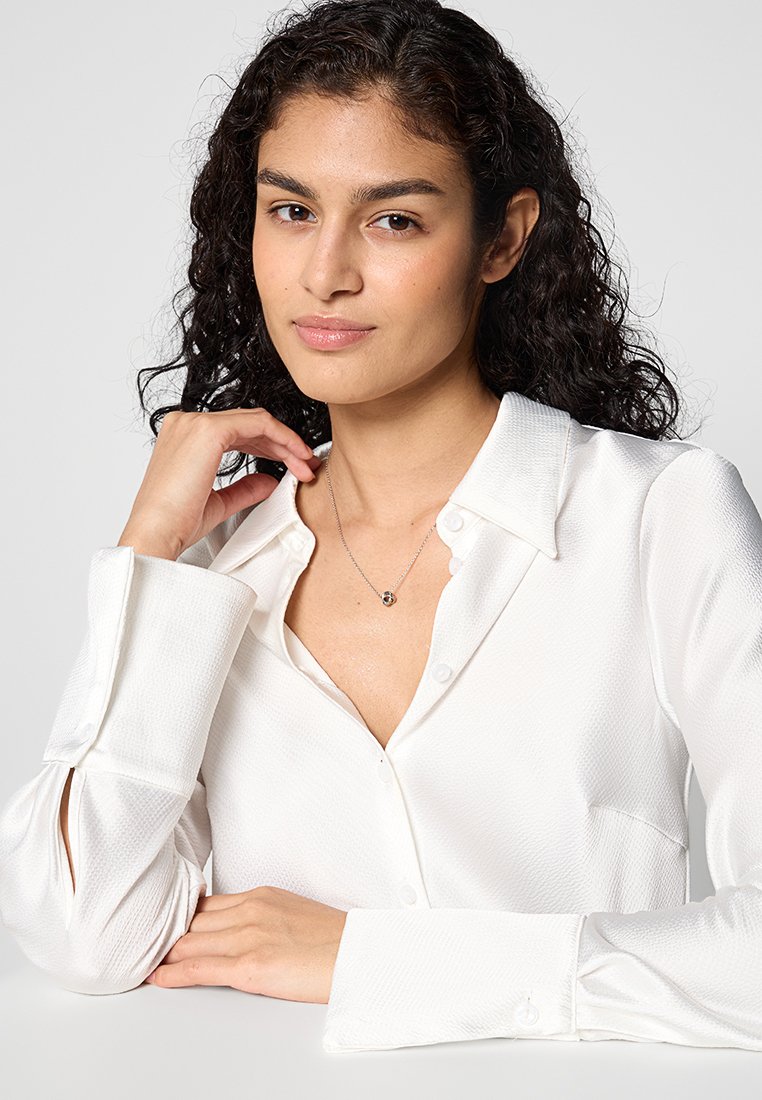 Witte blouse met een textuurfinish, voorzien van knoopsluitingen en opgerolde mouwen. Draagt een zilveren ketting.