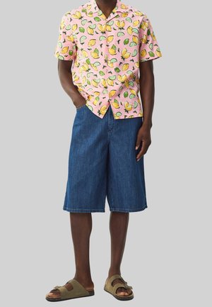 Homme portant une chemise rose à manches courtes avec un imprimé citron et lime, un short large en jean à la longueur genou et des sandales glissantes vert olive.