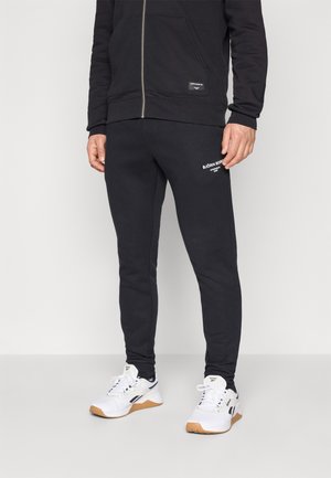 Schwarze Sweatpants aus Baumwollmischung mit schmalem Schnitt und weißem Logodetail am Oberschenkel, kombiniert mit weißen Sportschuhen.
