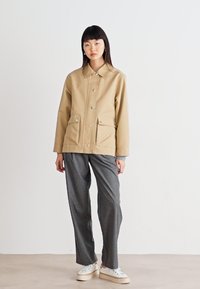 Beige, button-up jacket with two front pockets and a point collar. Paired with gray, loose-fit trousers and light sneakers. Smooth texture.